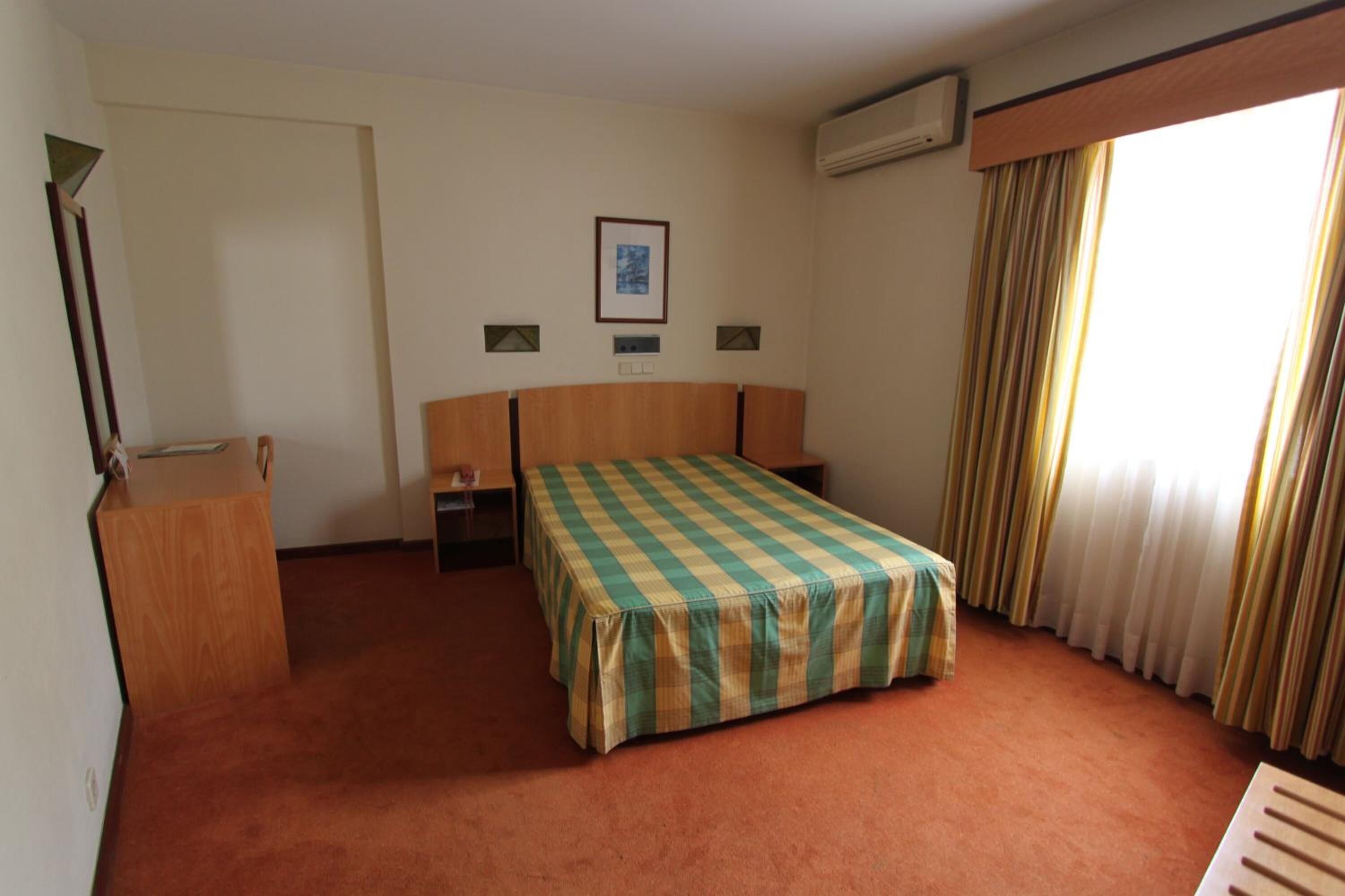 Une chambre d’hôtel avec un lit simple, une table de chevet en bois, un bureau et une fenêtre avec des rideaux.