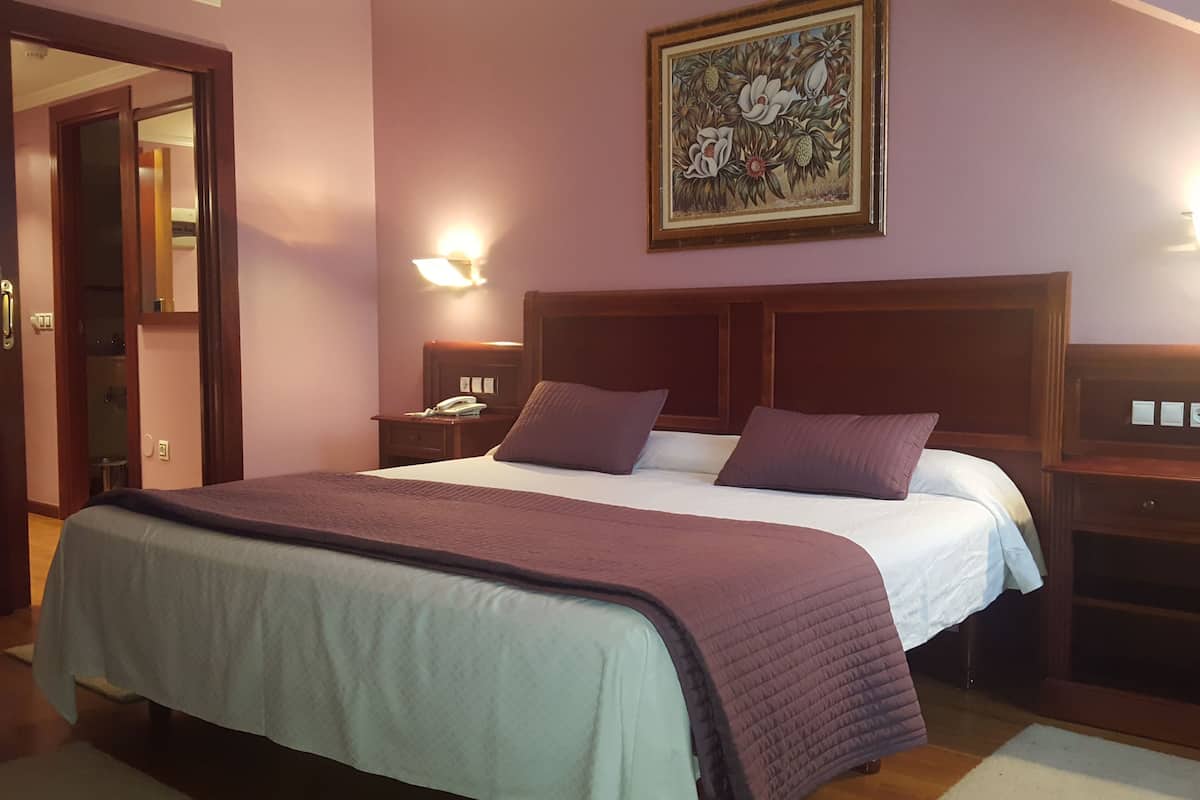 A hotel room with a large bed, two bedside tables, a nightstand, a painting on the wall, and a door leading to another room.
