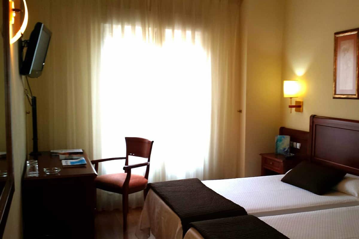 A hotel room with two beds, a desk with a computer, a chair, a lamp, and artwork on the wall.