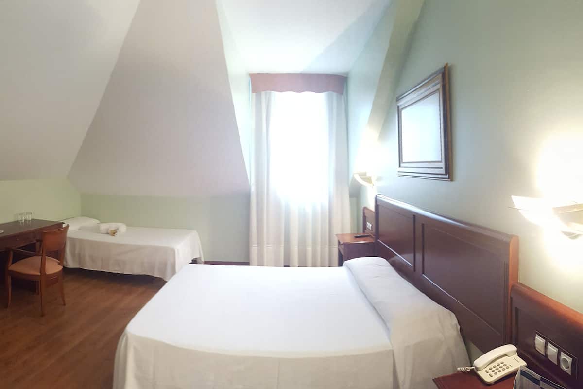 A hotel room with a large bed, a desk with a chair, two smaller beds, a telephone, and a window with curtains.