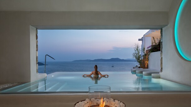 Une piscine à débordement moderne, une femme en méditation, une cheminée en verre et une vue sur la mer.