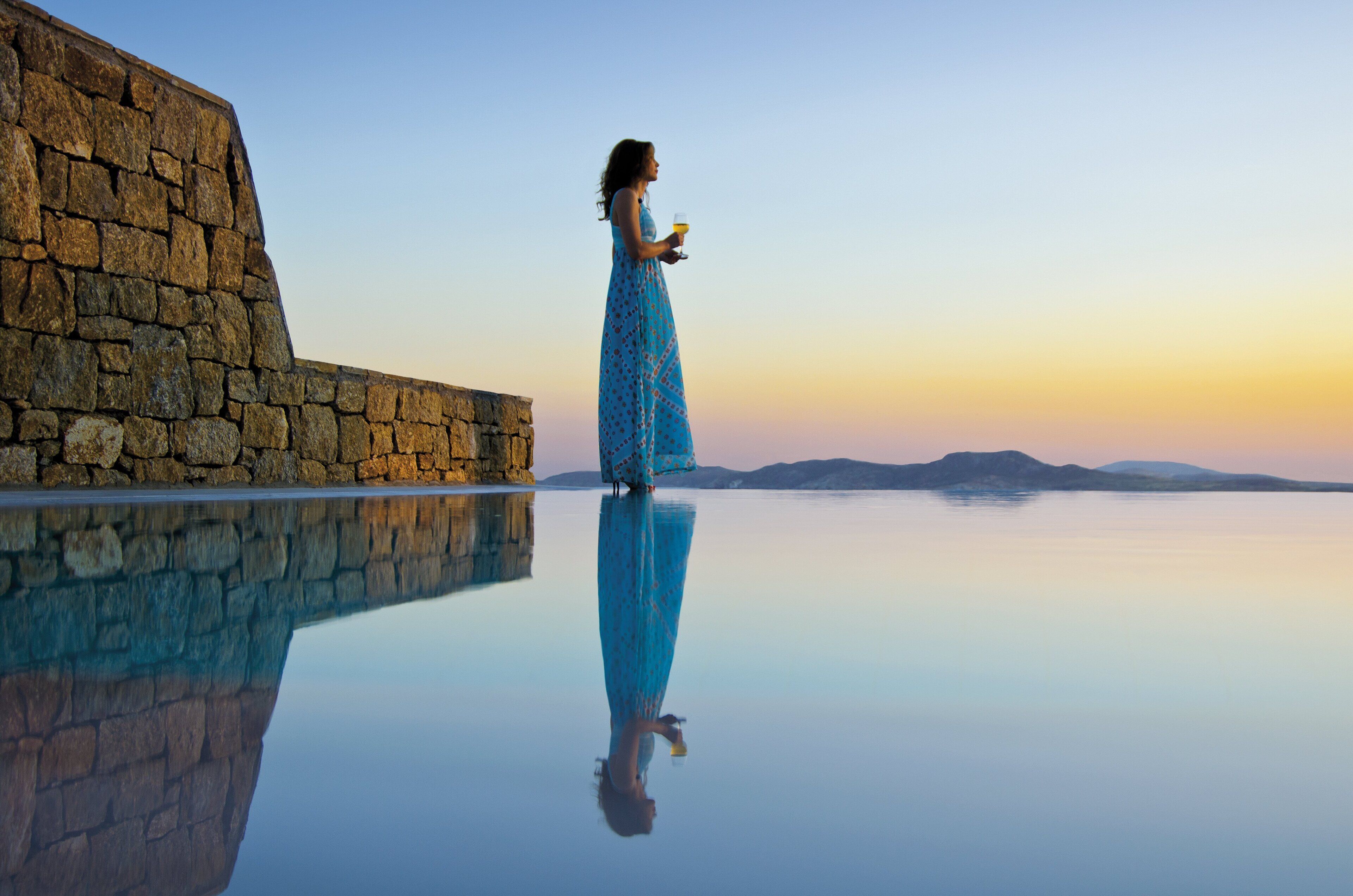 Una mujer con vestido azul parada junto a un muro de piedra, con un cuerpo de agua tranquilo que refleja su imagen.