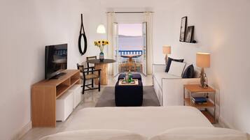 A hotel room with a bed, a desk with a chair, a small table with a vase of sunflowers, a sofa, a TV, and a view of the sea through an open door.