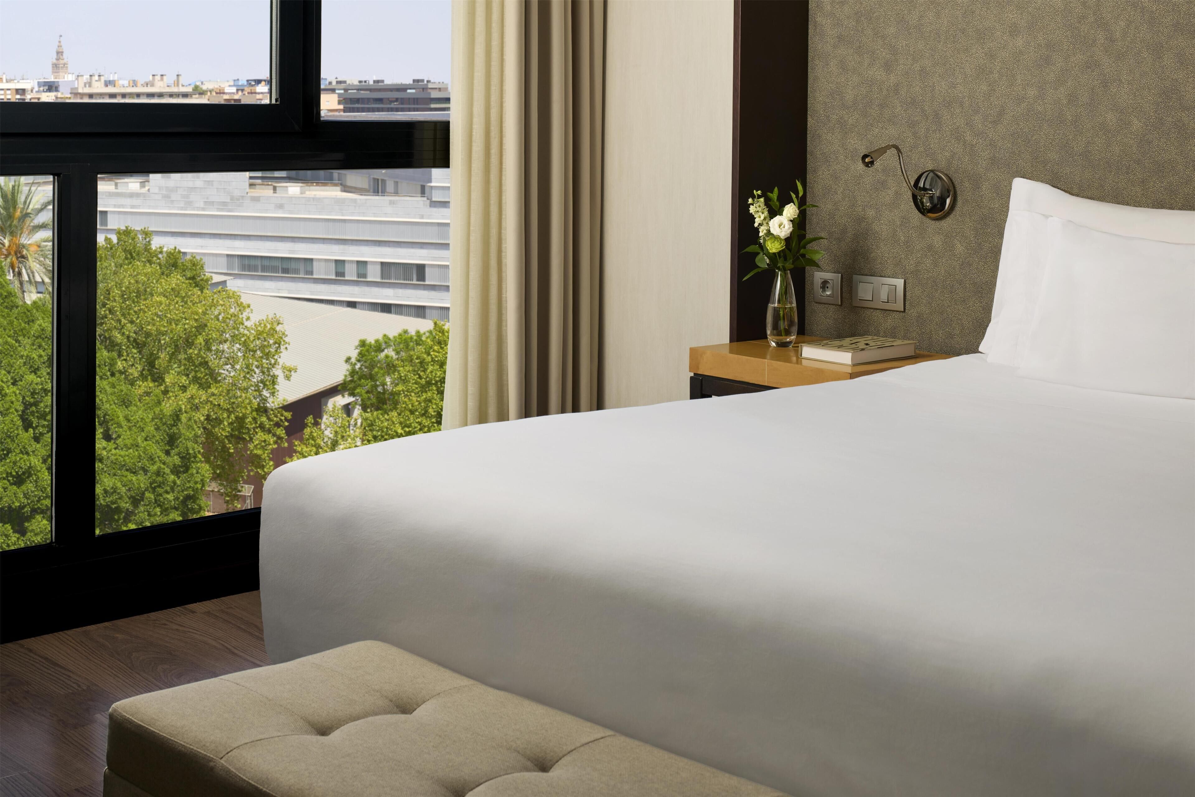 A hotel room with a large bed, a nightstand with a vase of flowers, a view of the cityscape, and a window with curtains.