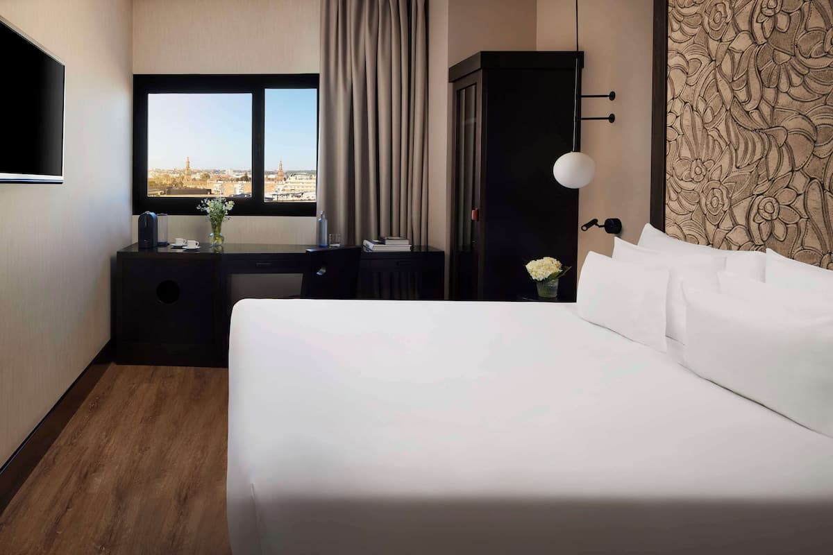 A hotel room with a large bed, a desk with a view of the city, a wardrobe, and a wall-mounted TV.