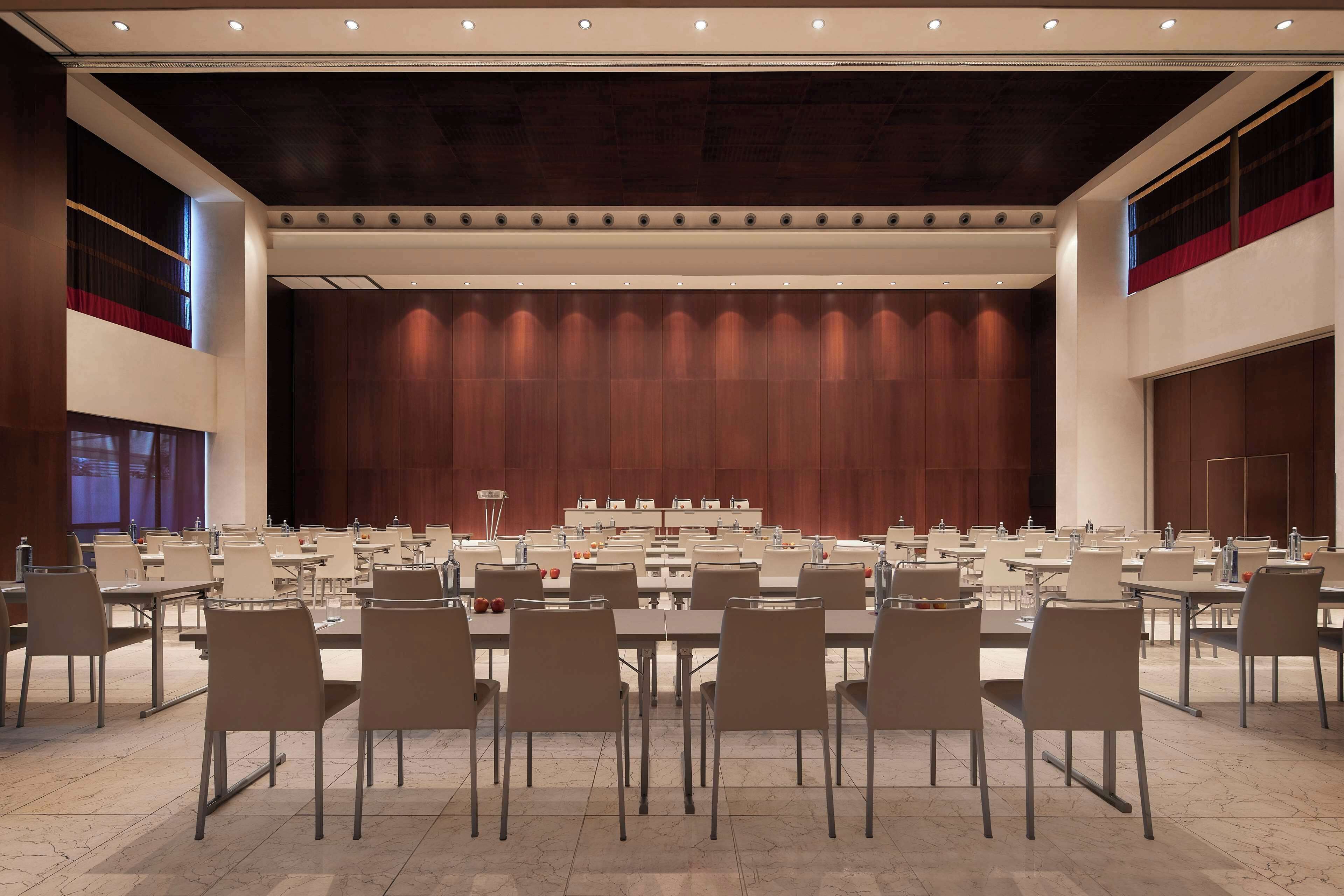 A conference room with multiple tables and chairs, a stage with a wooden backdrop, and a high ceiling with recessed lighting.