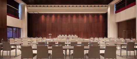 Une salle de conférence dotée de plusieurs tables et de chaises, d’une scène avec un décor en bois et d’un plafond haut avec des luminaires encastrés.