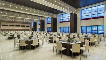 A large banquet hall with multiple round tables set with white tablecloths and chairs, large windows with blue curtains, and a high ceiling with recessed lighting.