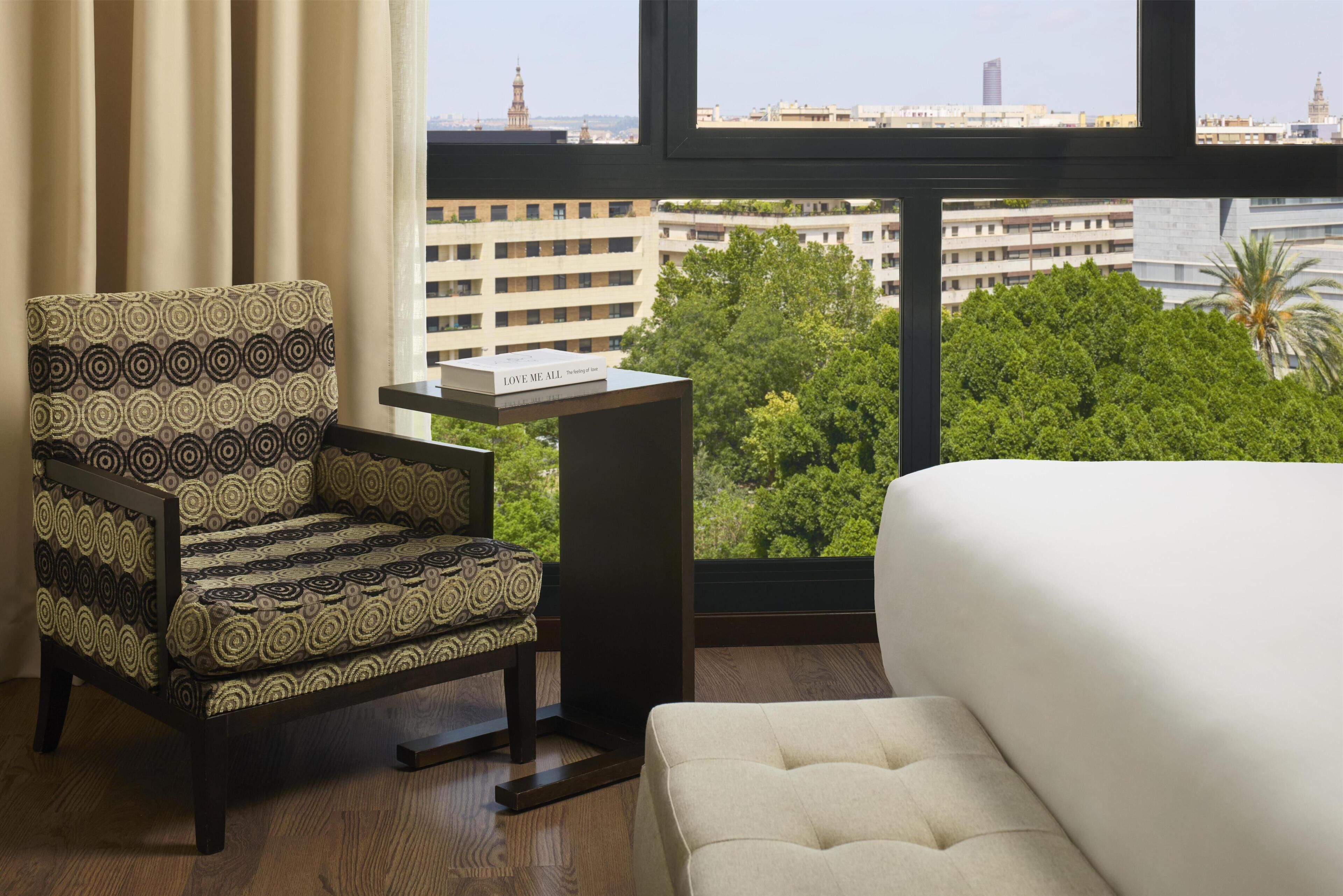 A hotel room with a patterned armchair, a small table, a white sofa, and a view of a cityscape through large windows.