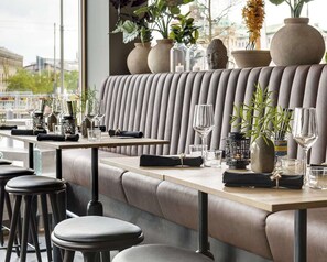 Un restaurante moderno con una amplia zona de asientos tipo banco, mesas con cristalería y plantas decorativas.
