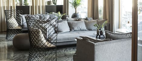A modern living room with a grey sofa, a patterned chair, and a coffee table. There are plants and a view of the city through large windows.