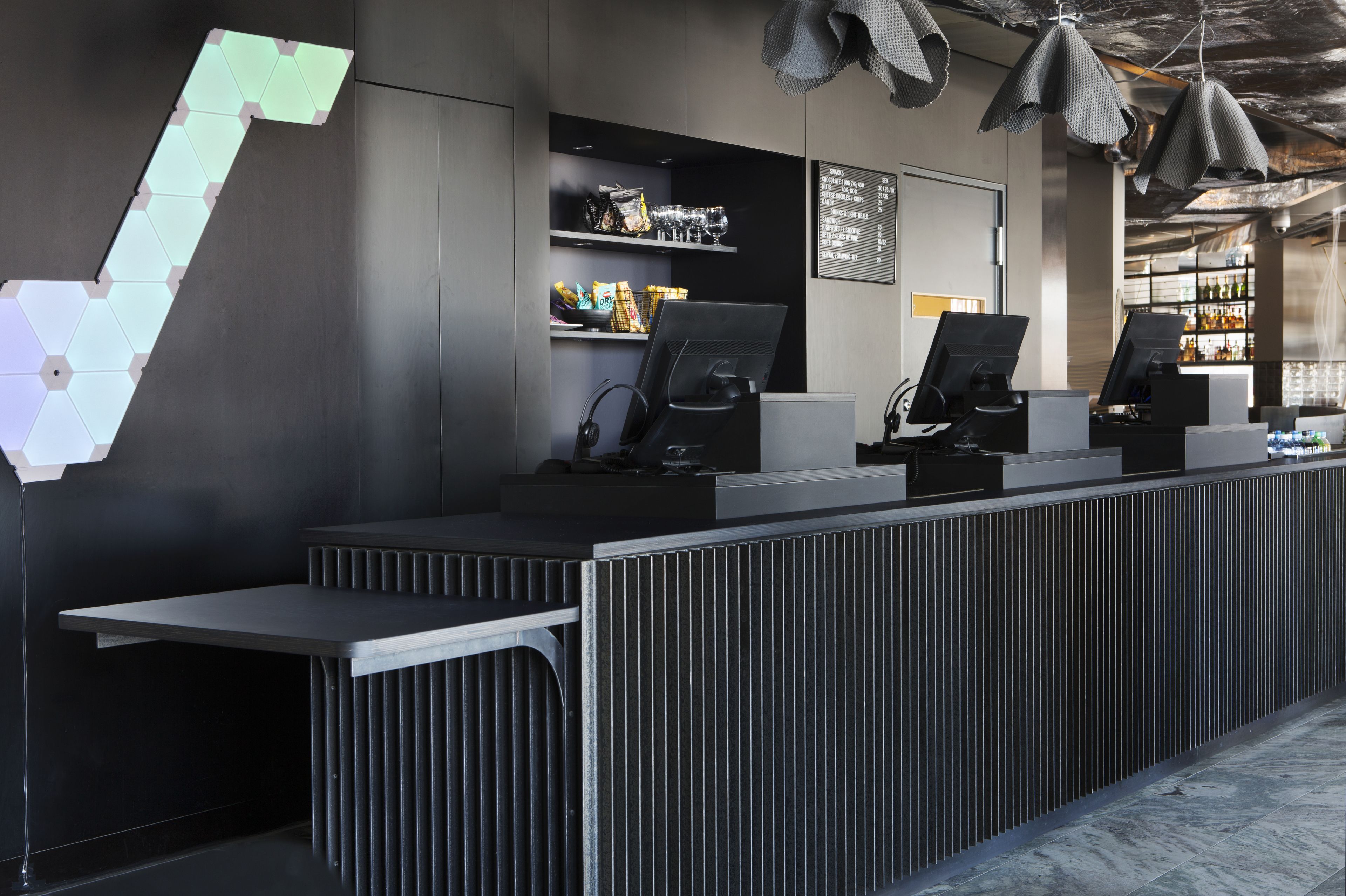 Un mostrador de recepción moderno con varias estaciones de trabajo de ordenador, un panel de menú y una zona de bar al fondo.