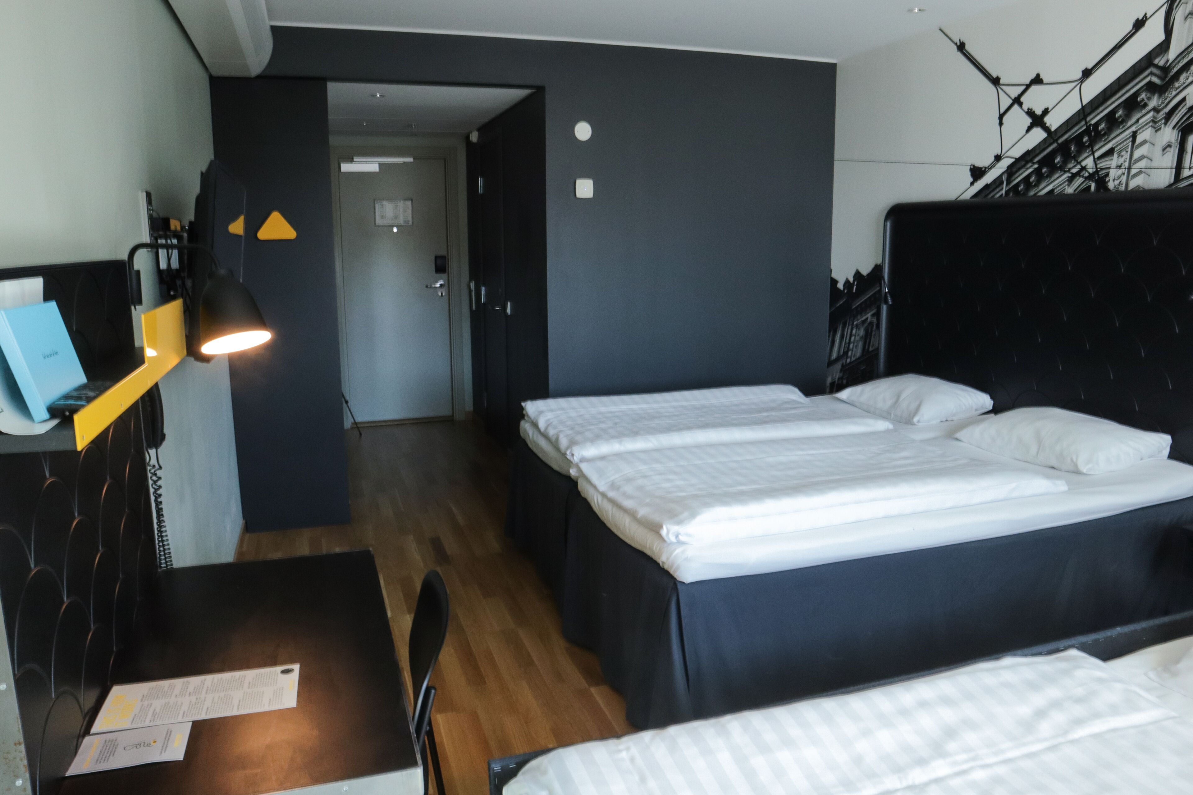 Una habitación de hotel con cama, escritorio y lámpara. Se ve una puerta que da a otra habitación.