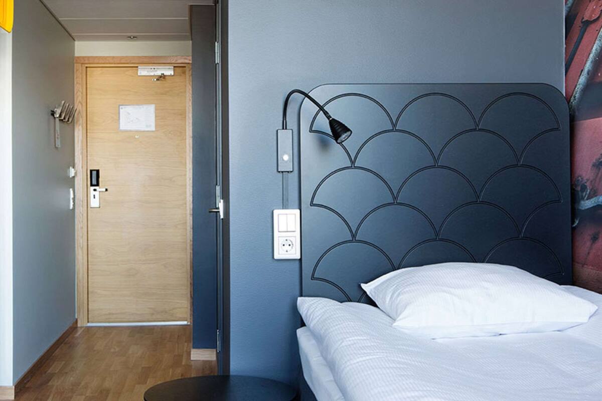 Una habitación de hotel con cama, mesita de noche, lámpara de pared y puerta con interruptor de luz.