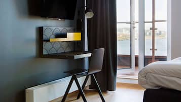Una habitación de hotel moderna con una cama, un escritorio con una silla, un televisor montado en la pared y vistas a una masa de agua a través de amplias ventanas.