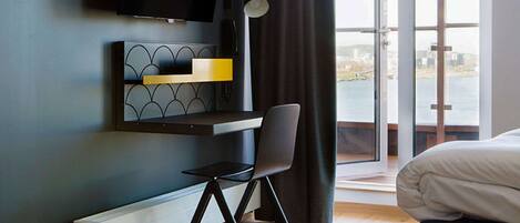 Una habitación de hotel moderna con una cama, un escritorio con una silla, un televisor montado en la pared y vistas a una masa de agua a través de amplias ventanas.