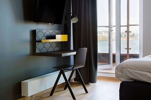 A modern hotel room with a bed, a desk with a chair, a TV mounted on the wall, and a view of a body of water through large windows.