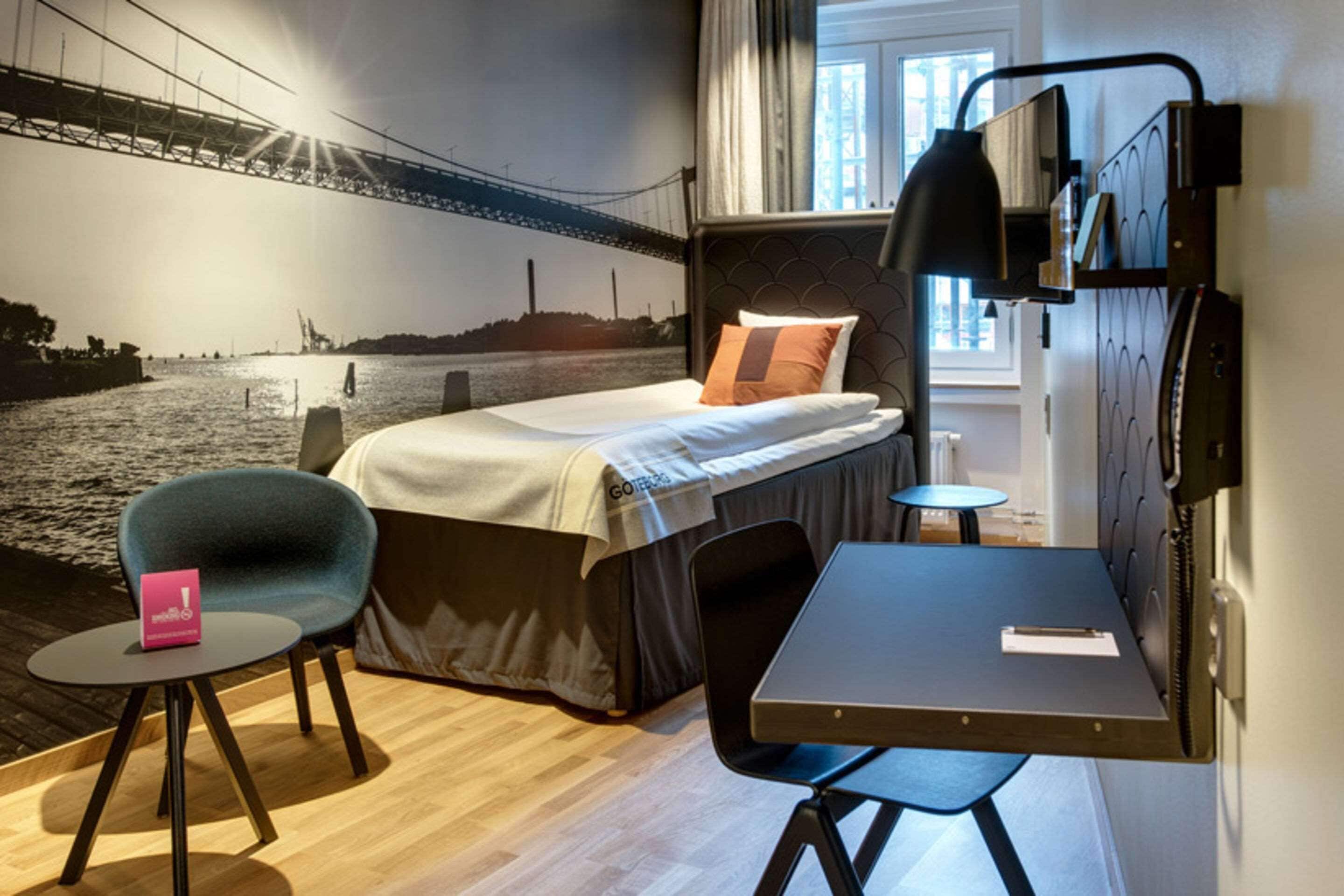 Ein Hotelzimmer mit Bett, Schreibtisch, Stuhl und Wandbild einer Brücke, sowie einem Fenster.