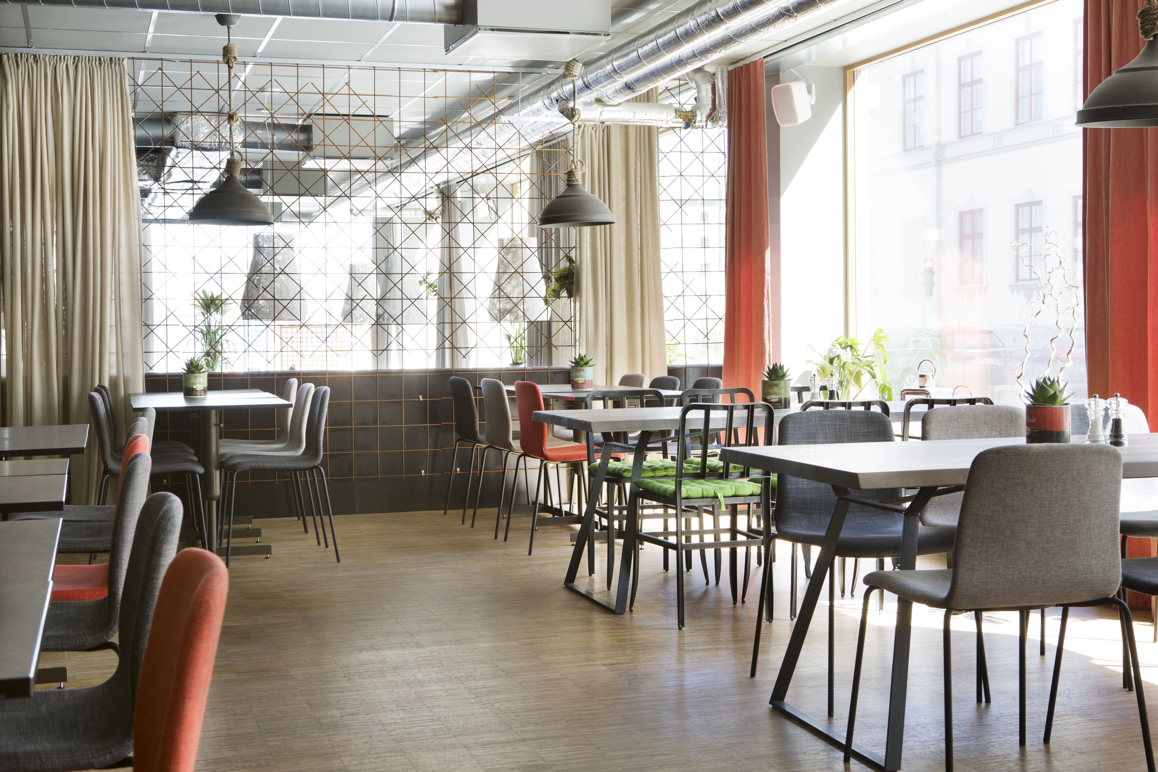 Un espace de restauration moderne avec des tables et des chaises, de grandes fenêtres avec des rideaux, et des éléments structurels apparents, comme des tuyaux et des poutres.