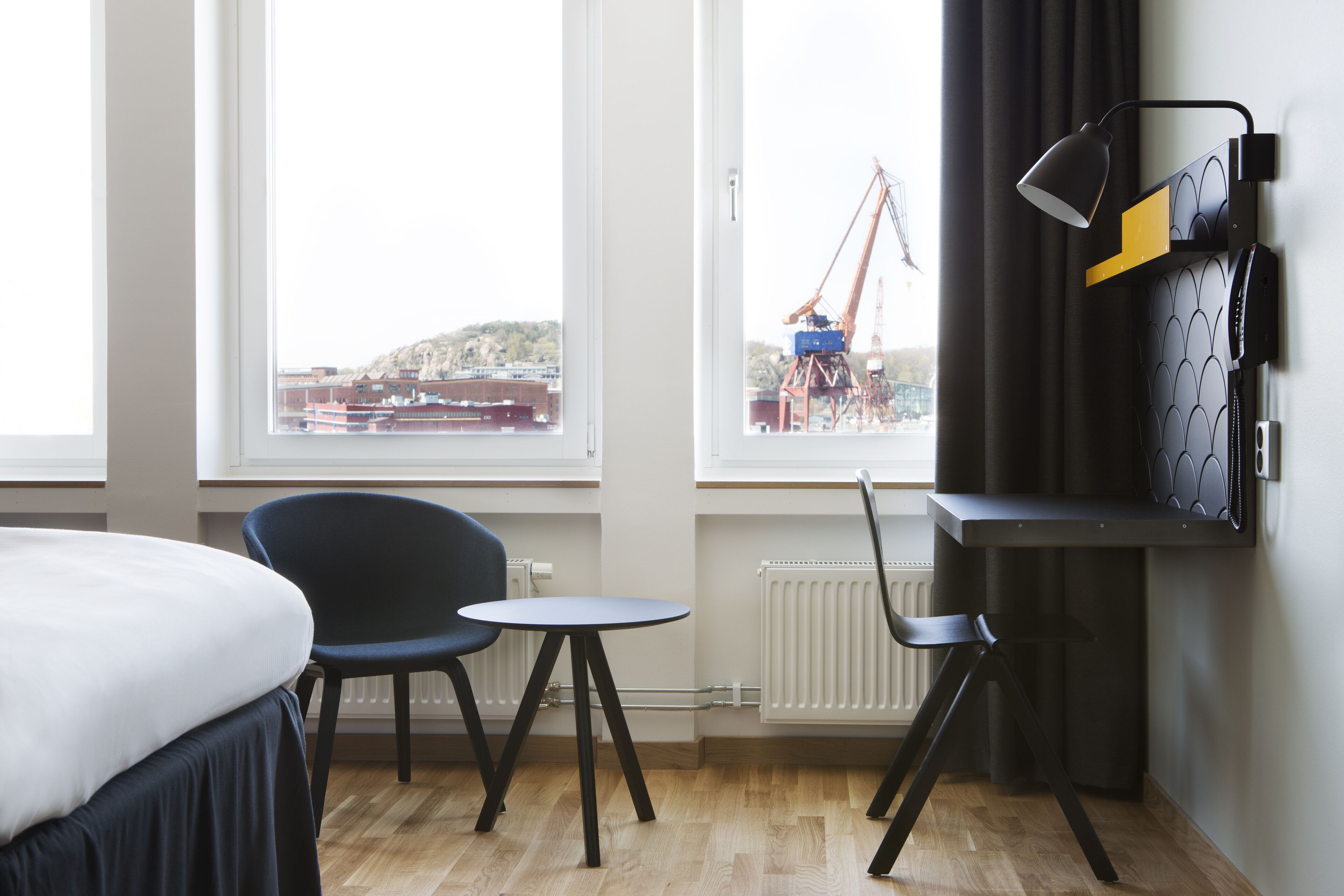 A hotel room with a bed, a desk, a chair, a table, a lamp, and a view of a construction site through the window.