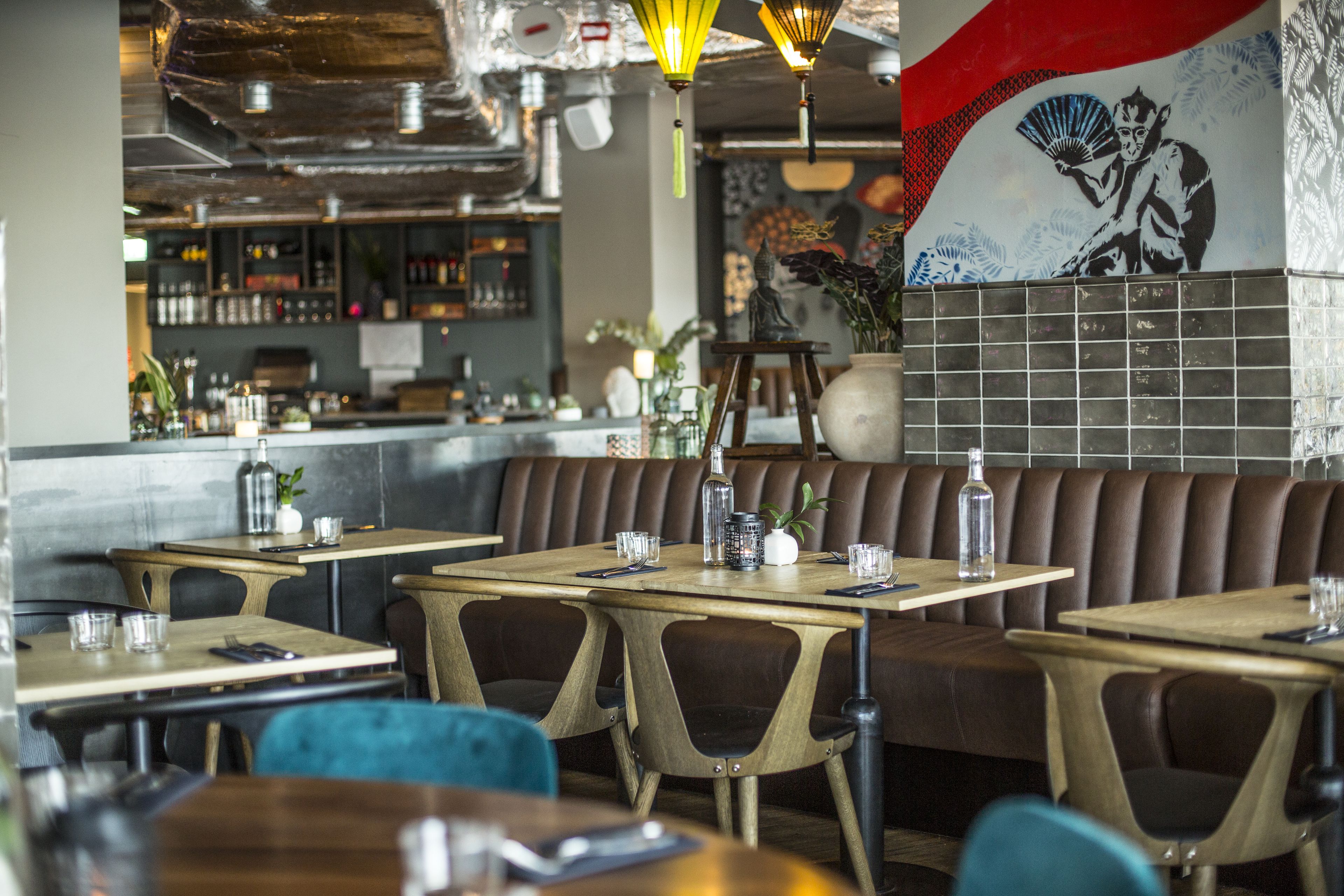 A restaurant interior with wooden tables and booths, a bar area in the background, and a decorative wall with a fan and floral pattern.
