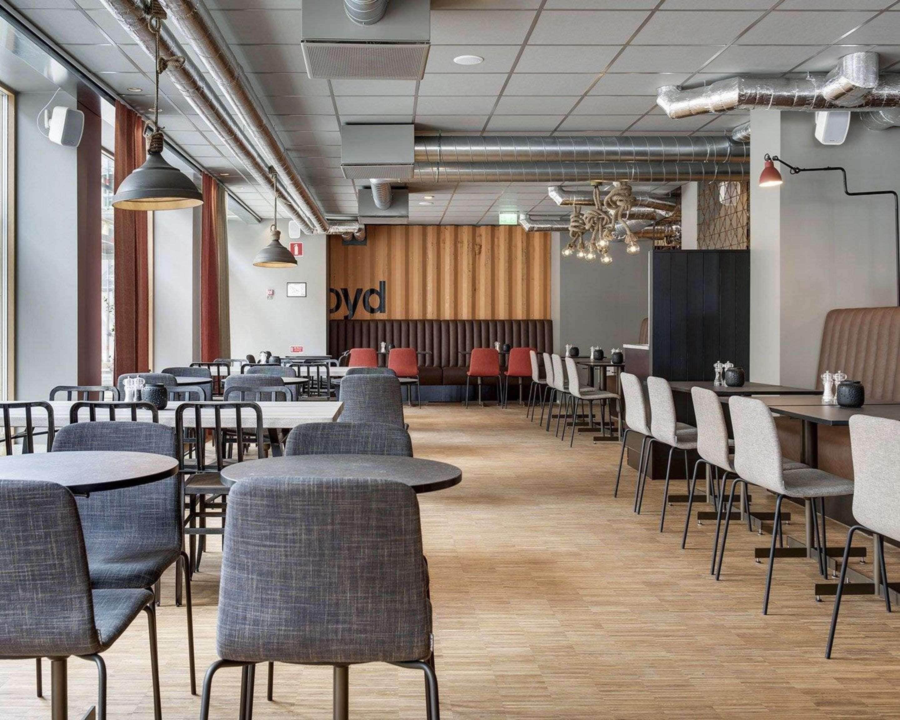 A modern restaurant with high ceilings, exposed ductwork, and a mix of round and rectangular tables with grey upholstered chairs.