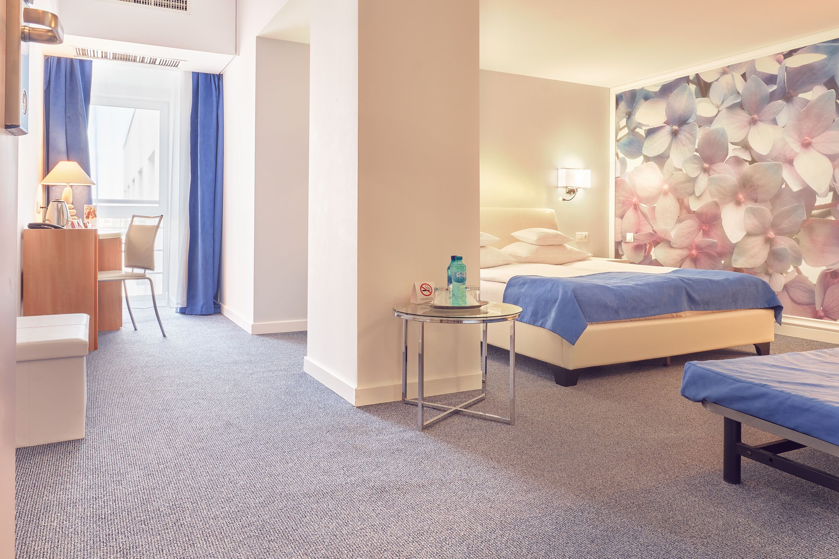A hotel room with a bed, a small table with a bottle, a chair, and a wall with a floral pattern.