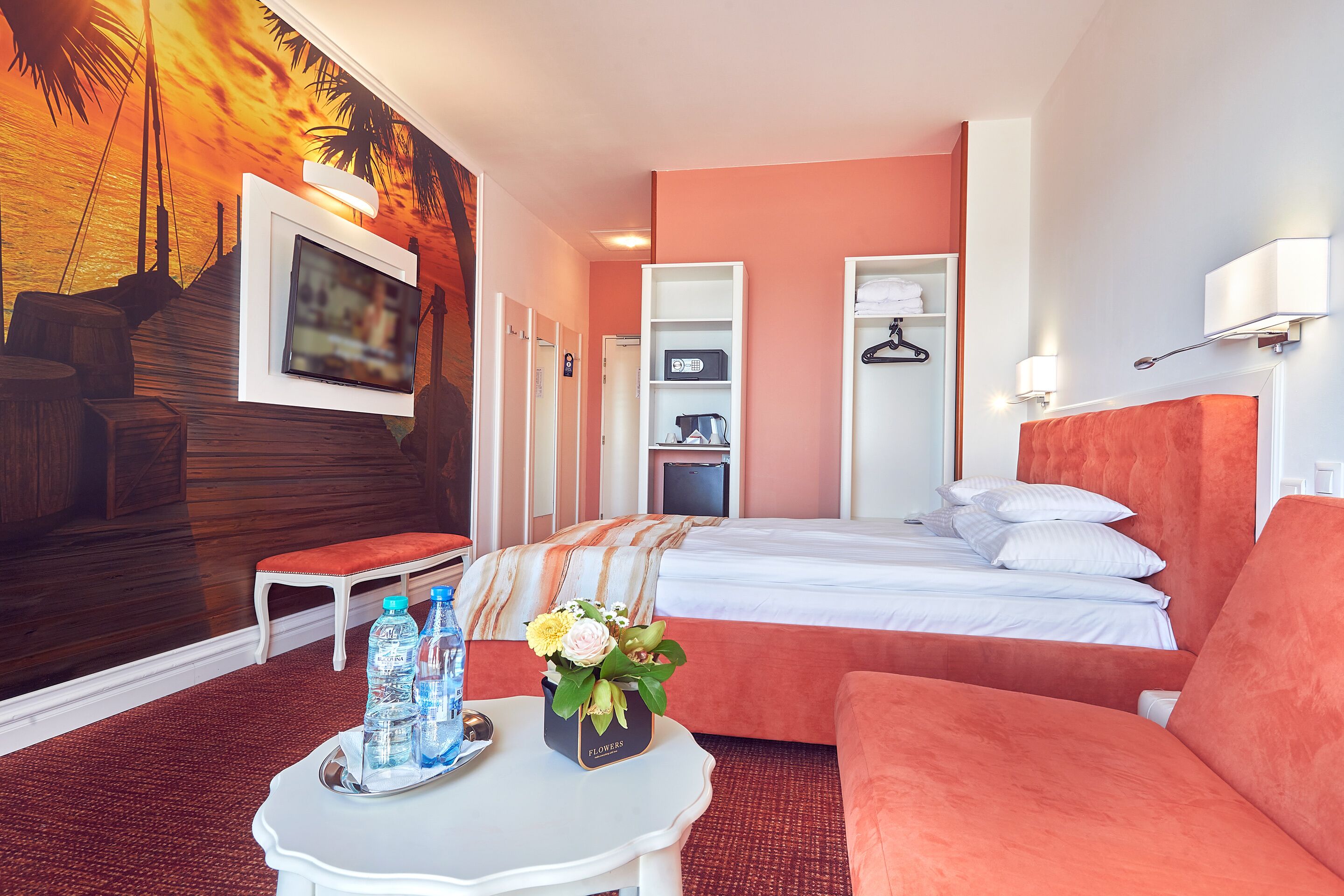 Habitación de hotel con cama, sofá, televisión, estante montado en la pared y un mural de atardecer en la pared.