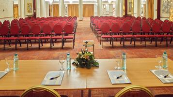 A conference room with rows of red chairs, tables with water bottles, glasses, and notepads.