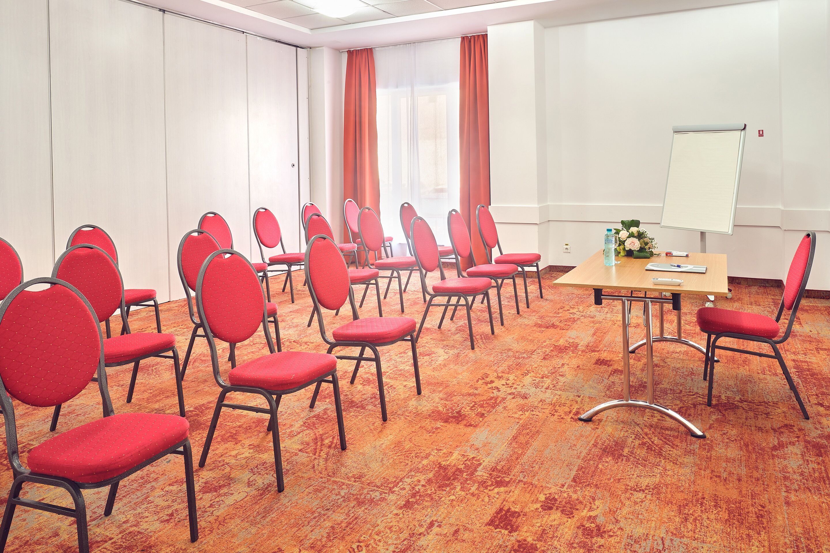 A conference room with red chairs arranged in rows, a wooden table, and a whiteboard.