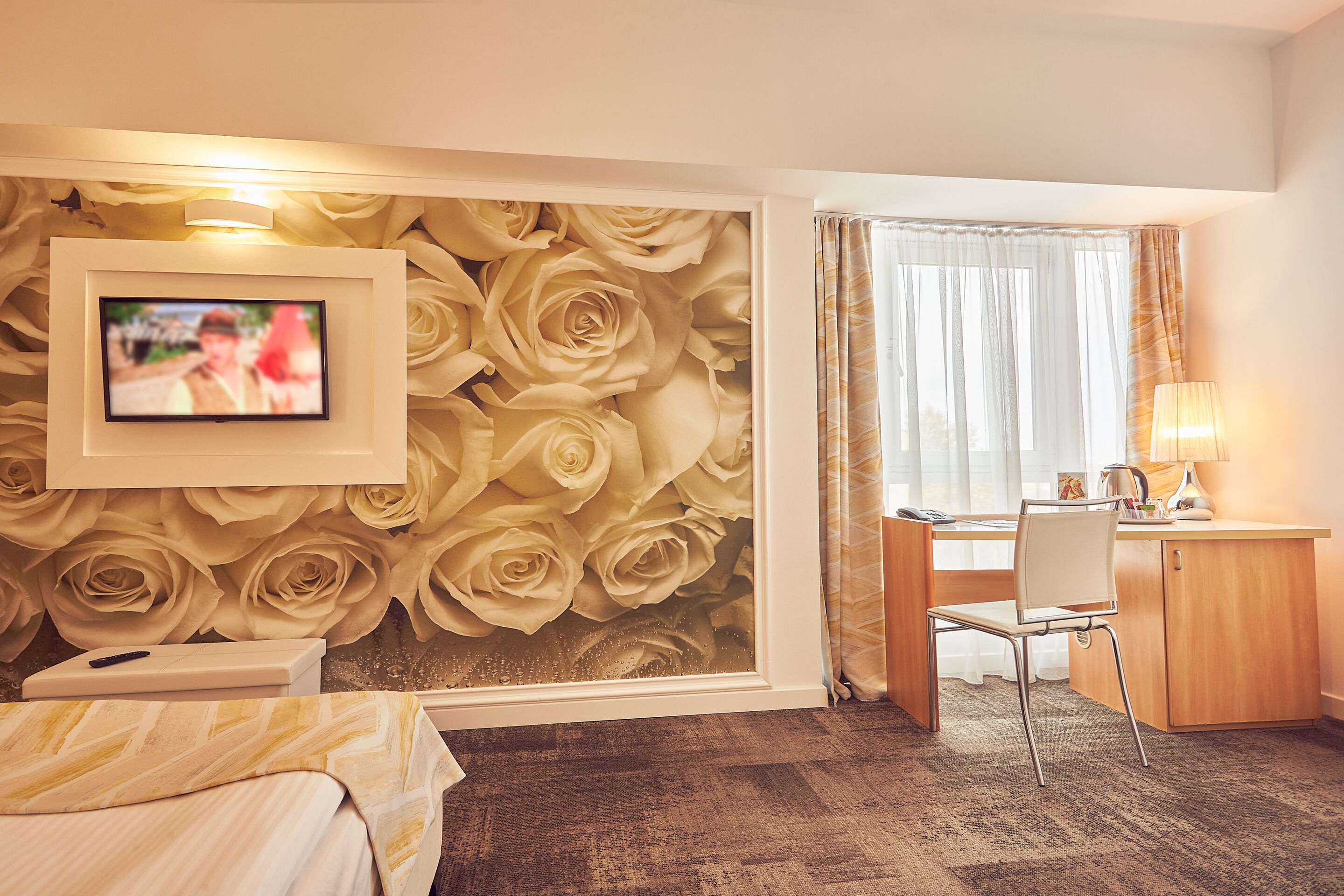 A hotel room with a large wall mural of roses, a desk with a chair, a bed with a striped bedspread, and a window with sheer curtains.