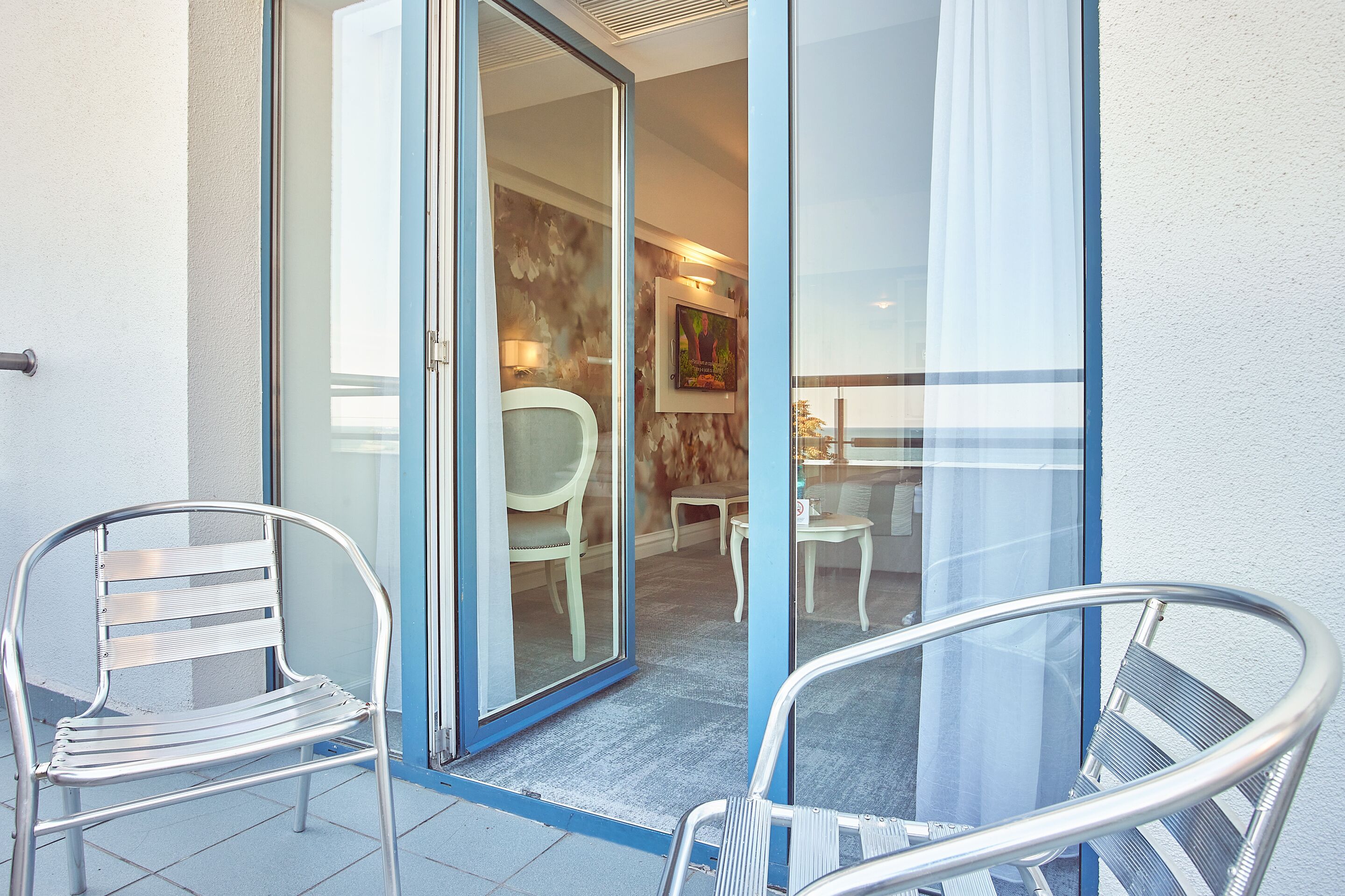 A balcony with two metal chairs, a glass door leading inside, and a view of the interior including a dining area and a painting on the wall.