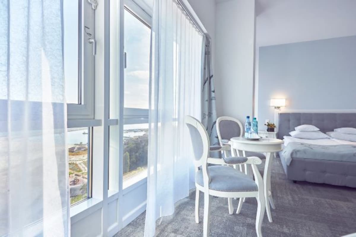 Quarto de hotel com uma janela grande, uma cama, uma mesinha de cabeceira com garrafas de água e uma cadeira.