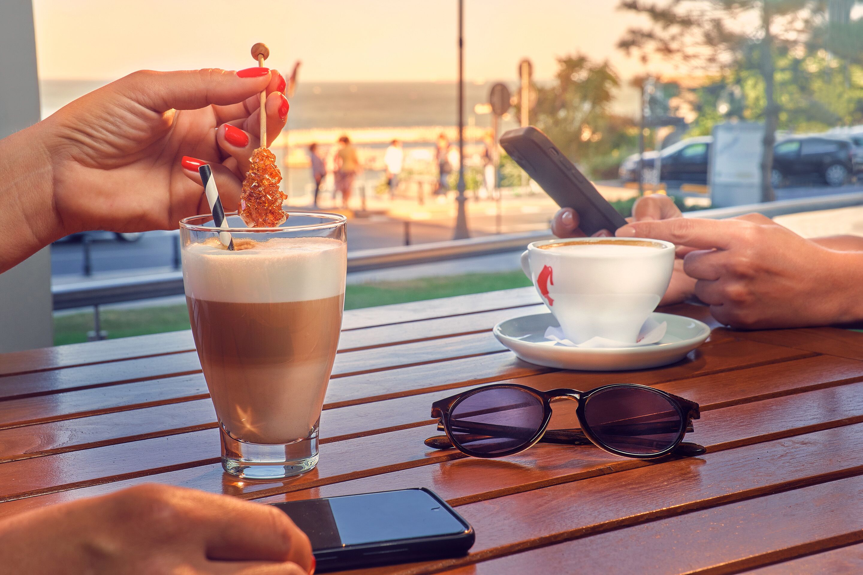 A wooden table with a glass of iced coffee, a cup of coffee, sunglasses, and a smartphone.