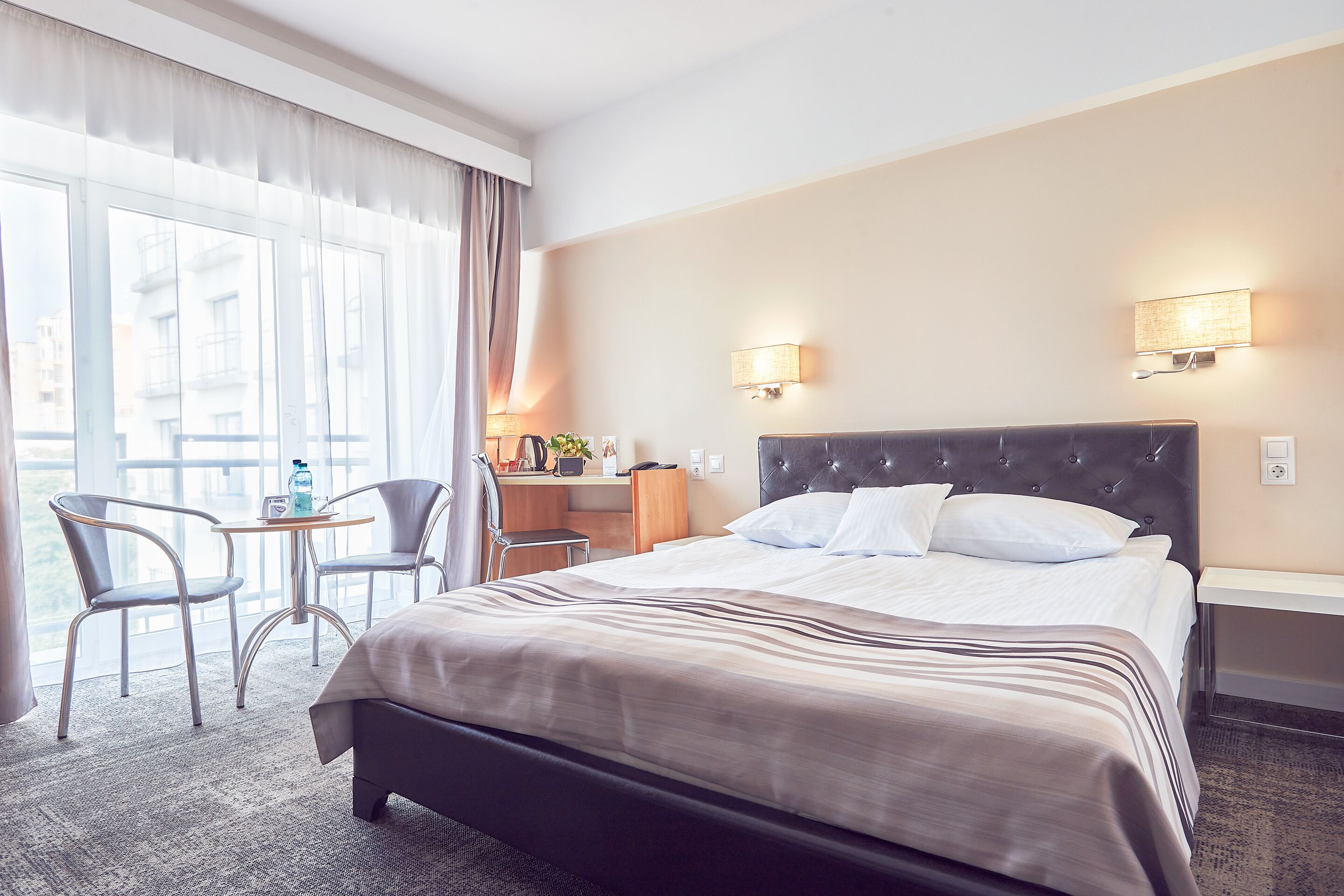 Een hotelkamer met een groot bed, twee nachtkastjes, een bureau met een stoel, en uitzicht door grote ramen.