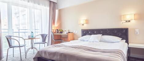 A hotel room with a large bed, two bedside tables, a desk with a chair, and a view through large windows.