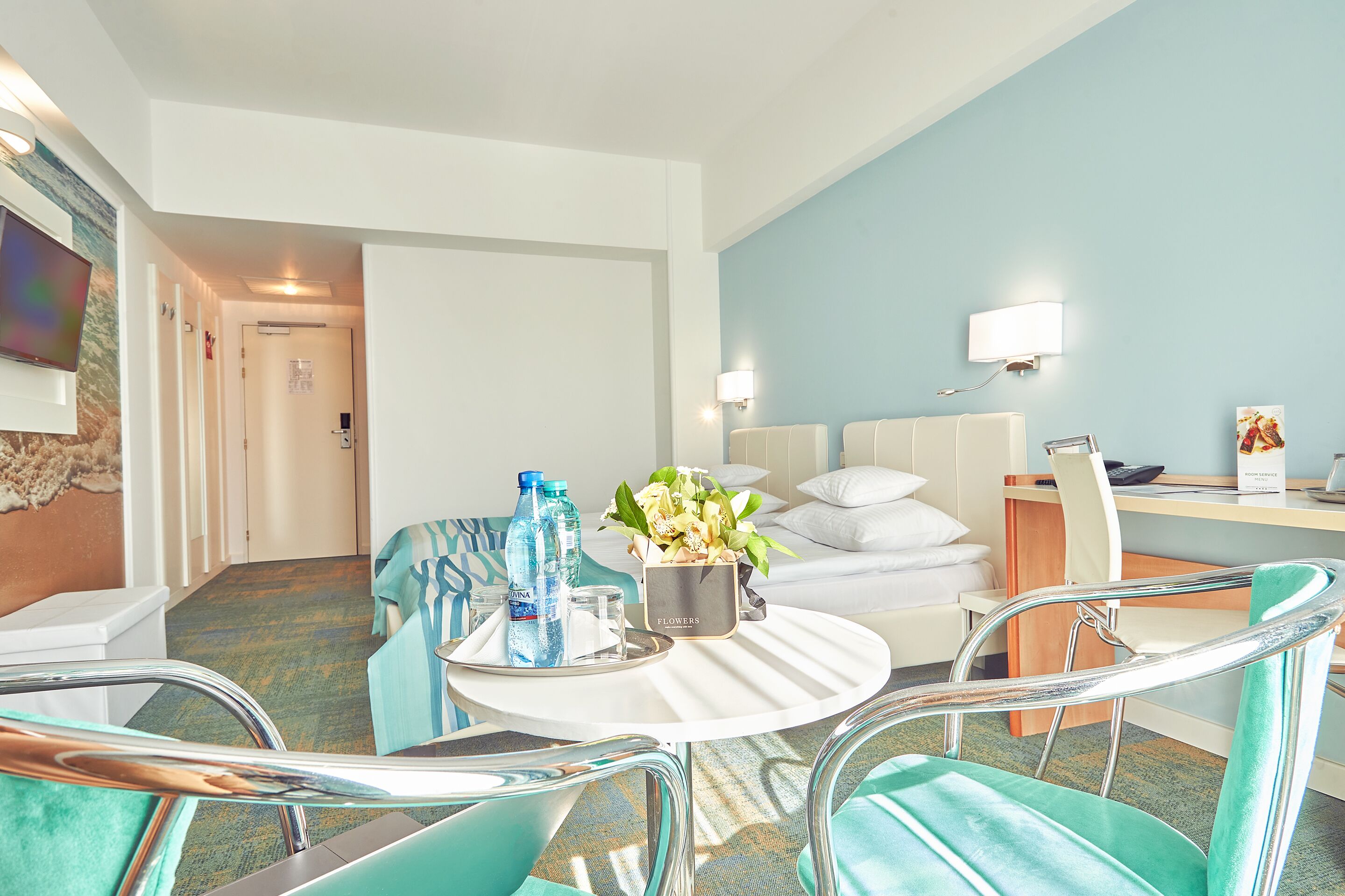 A hotel room with a bed, a desk, a chair, a round table with a tray of water bottles and a flower vase, and a television on the wall.