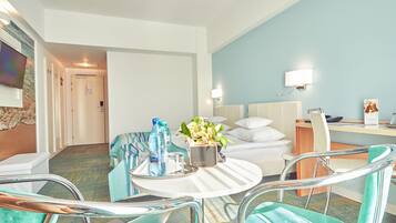 Una habitación de hotel con una cama, un escritorio, una silla, una mesa redonda con un recipiente de botellas de agua y un jarrón, y un televisor en la pared.