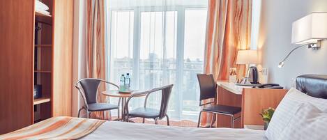 A hotel room with a large bed, two chairs, a small table, a desk, and a window with curtains.