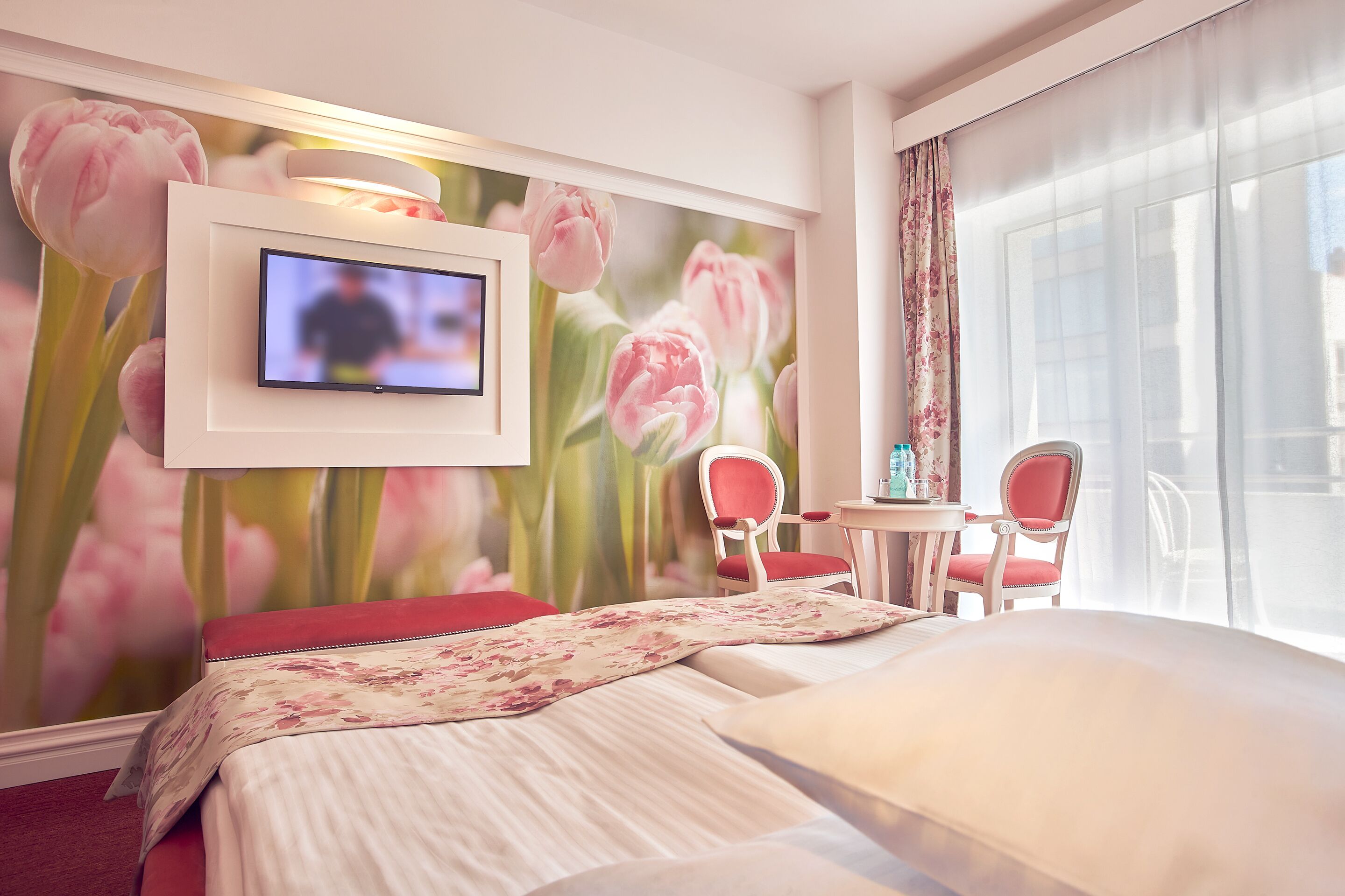 A hotel room with a bed, a TV mounted on the wall, a floral mural, a balcony with a table and chairs, and a large window.