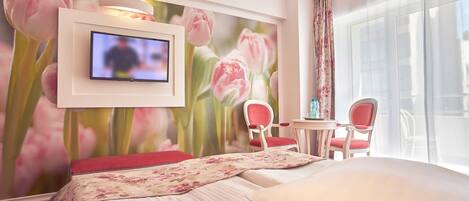 Habitación de hotel con cama, televisor montado en la pared, un mural floral, un balcón con mesa y sillas, y un ventanal grande.