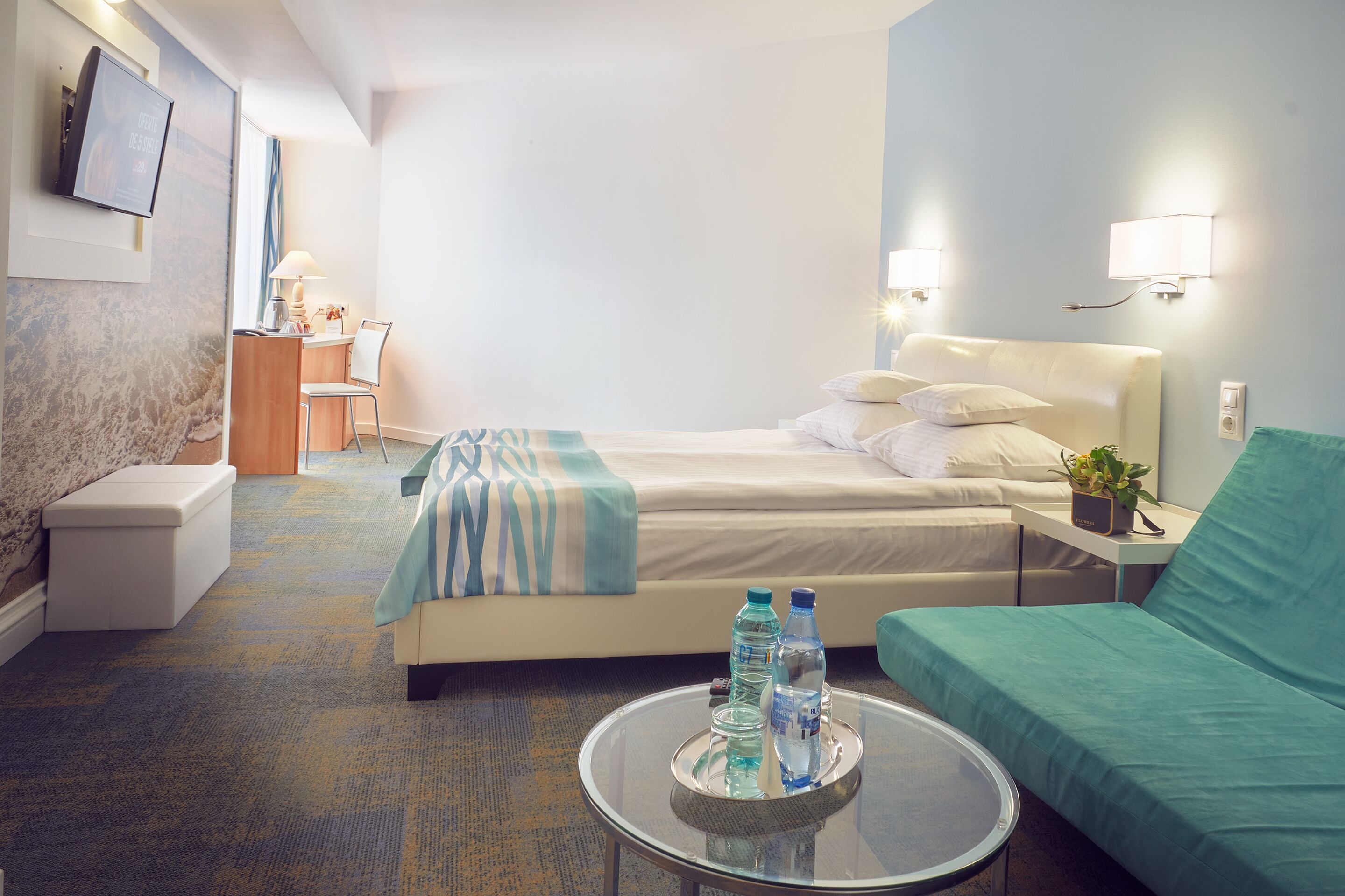 A hotel room with a bed, a green sofa, a small table with water bottles, a desk with a lamp, and a TV on the wall.