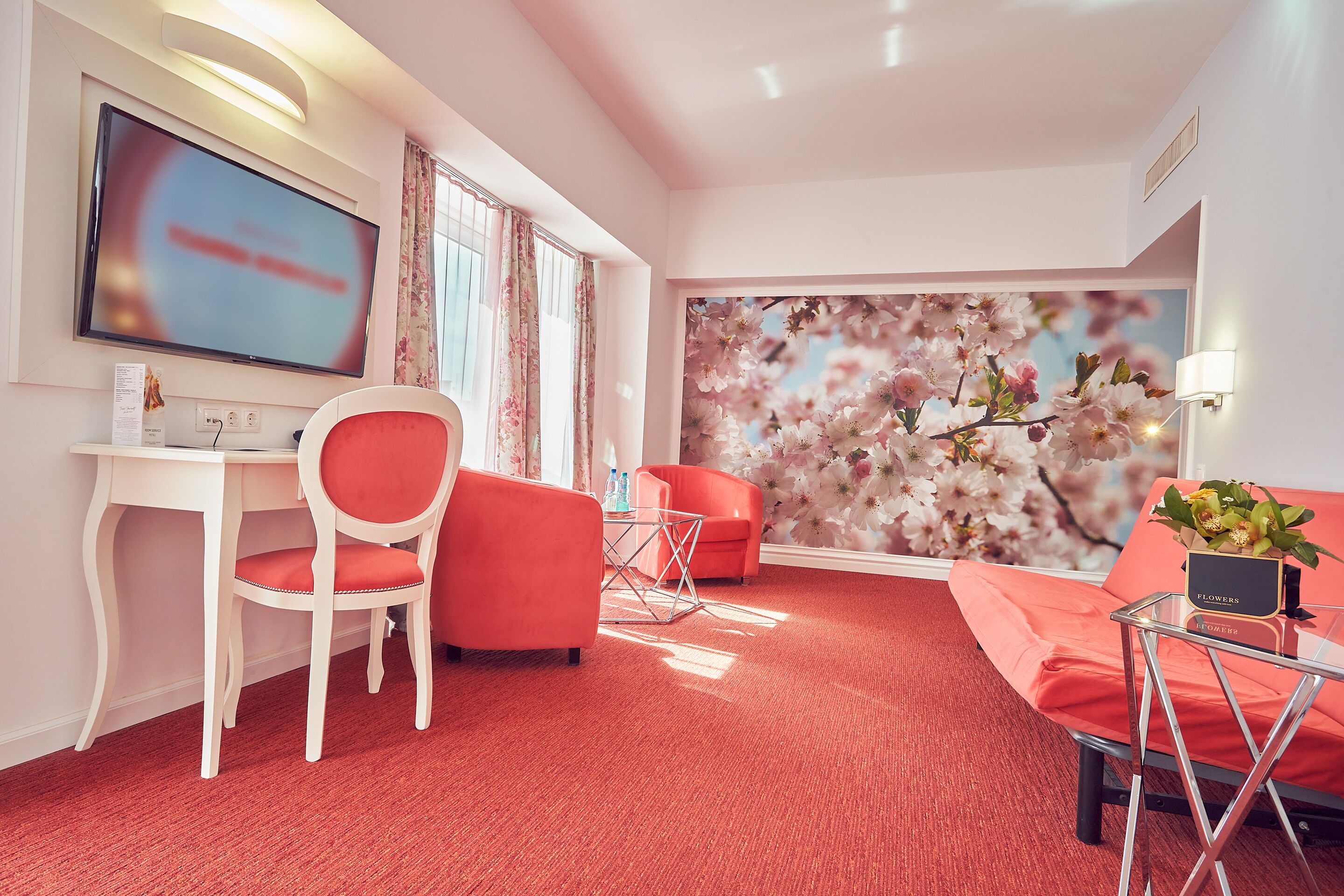 Una habitación de hotel moderna con alfombra roja, un escritorio blanco, una silla roja, un sofá rojo y un televisor montado en la pared.