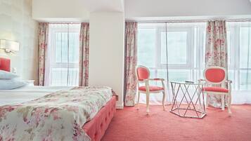 A hotel room with a bed, two chairs, a small table, and large windows with curtains.