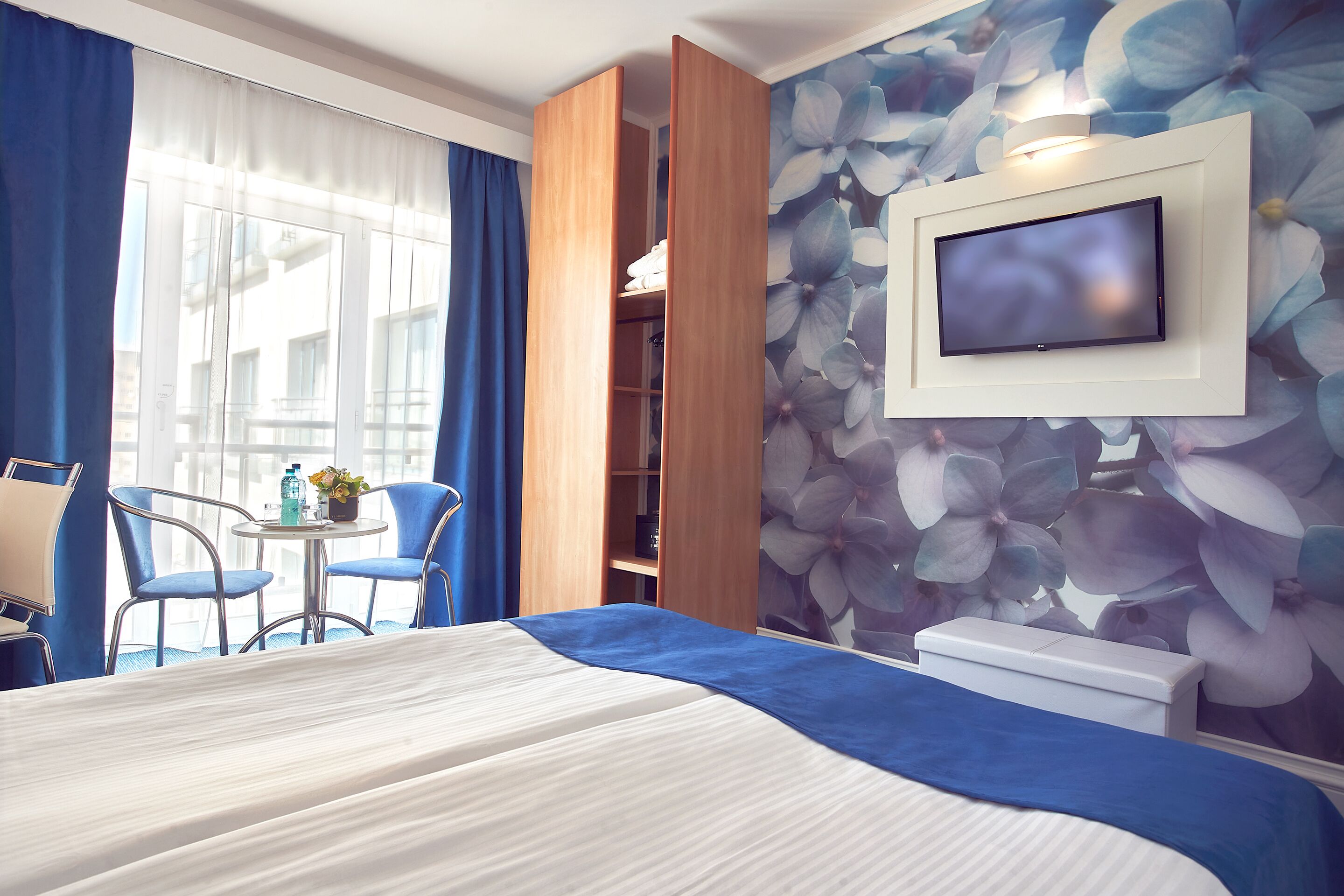 Una habitación de hotel con cama, televisor montado en la pared, una pequeña mesa con sillas y una ventana con cortinas.