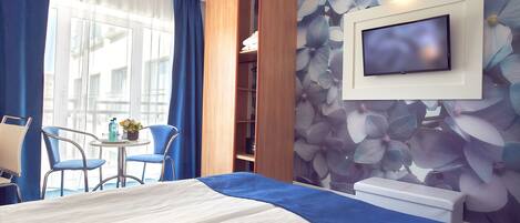 Una habitación de hotel con cama, televisor montado en la pared, una pequeña mesa con sillas y una ventana con cortinas.