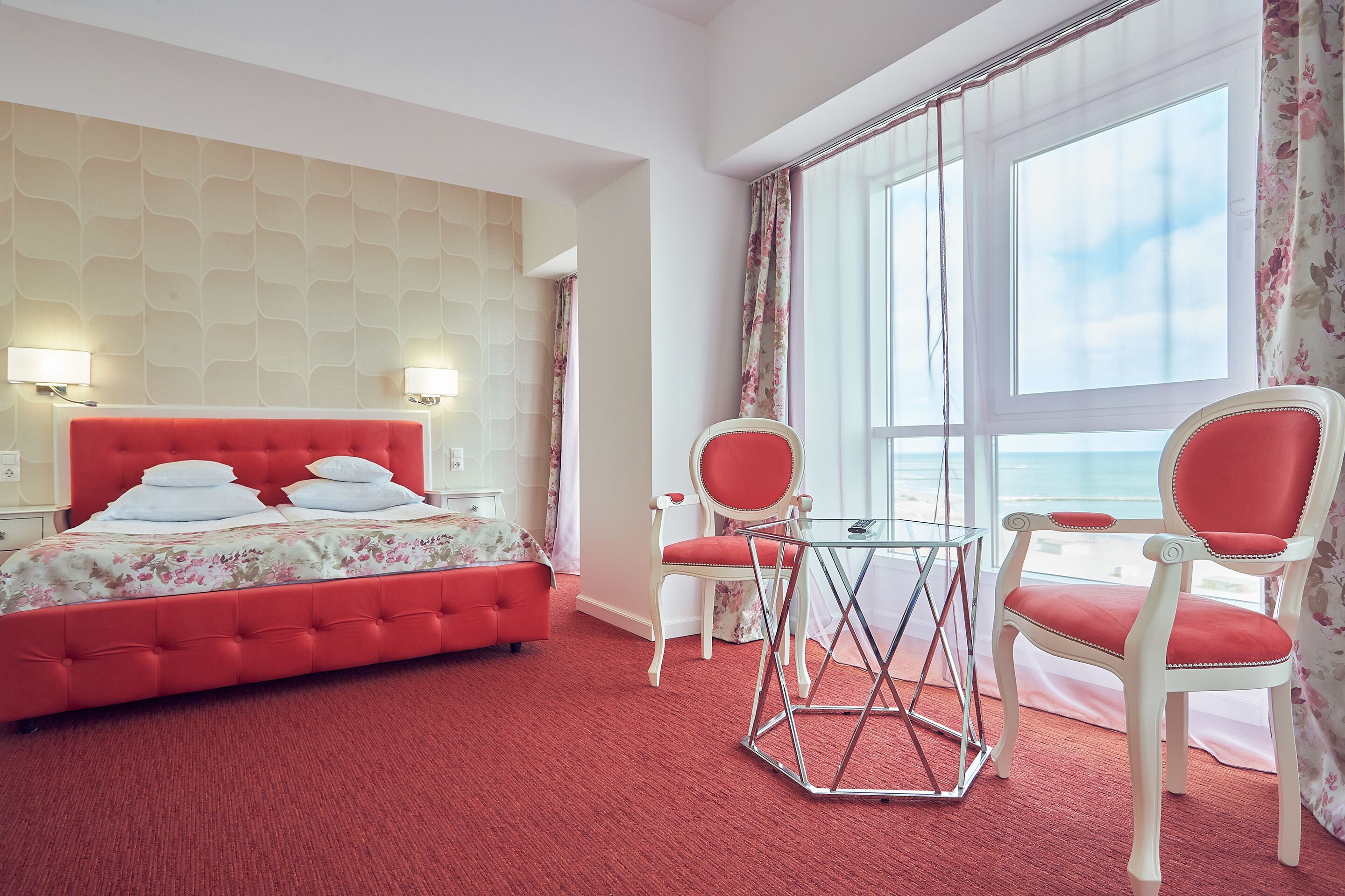 Une chambre d’hôtel avec un tapis rouge, un lit rouge et deux fauteuils rouges. Il y a une fenêtre avec des rideaux et une vue sur le ciel.