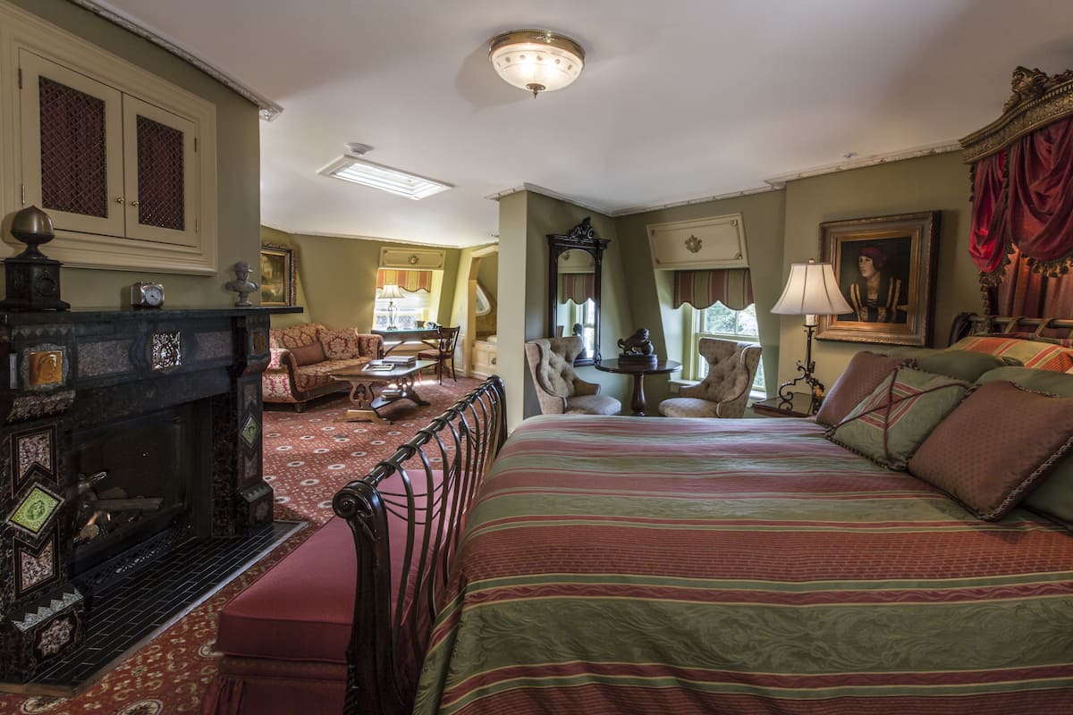 A traditional room with a fireplace, a bed with striped bedding, a dining area with a table and chairs, and a mirror on the wall.