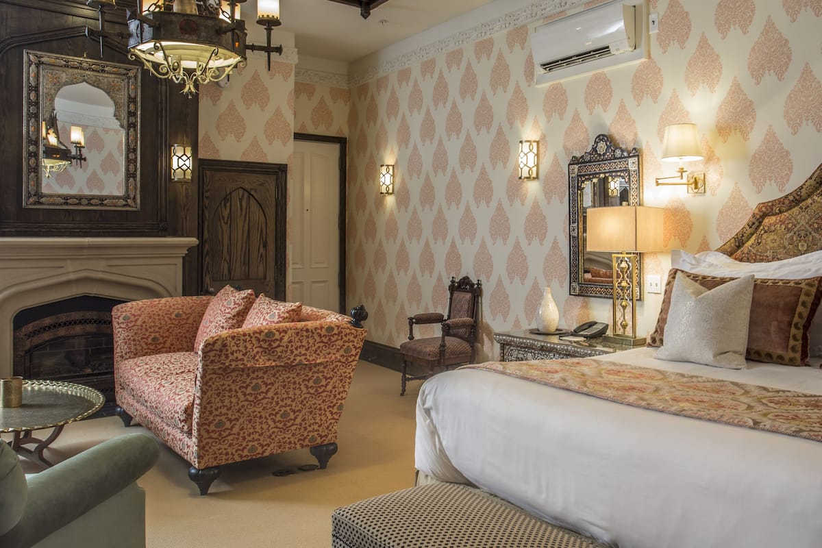 A hotel room with a large bed, a sitting area with a chair, a fireplace, and decorative wall sconces.