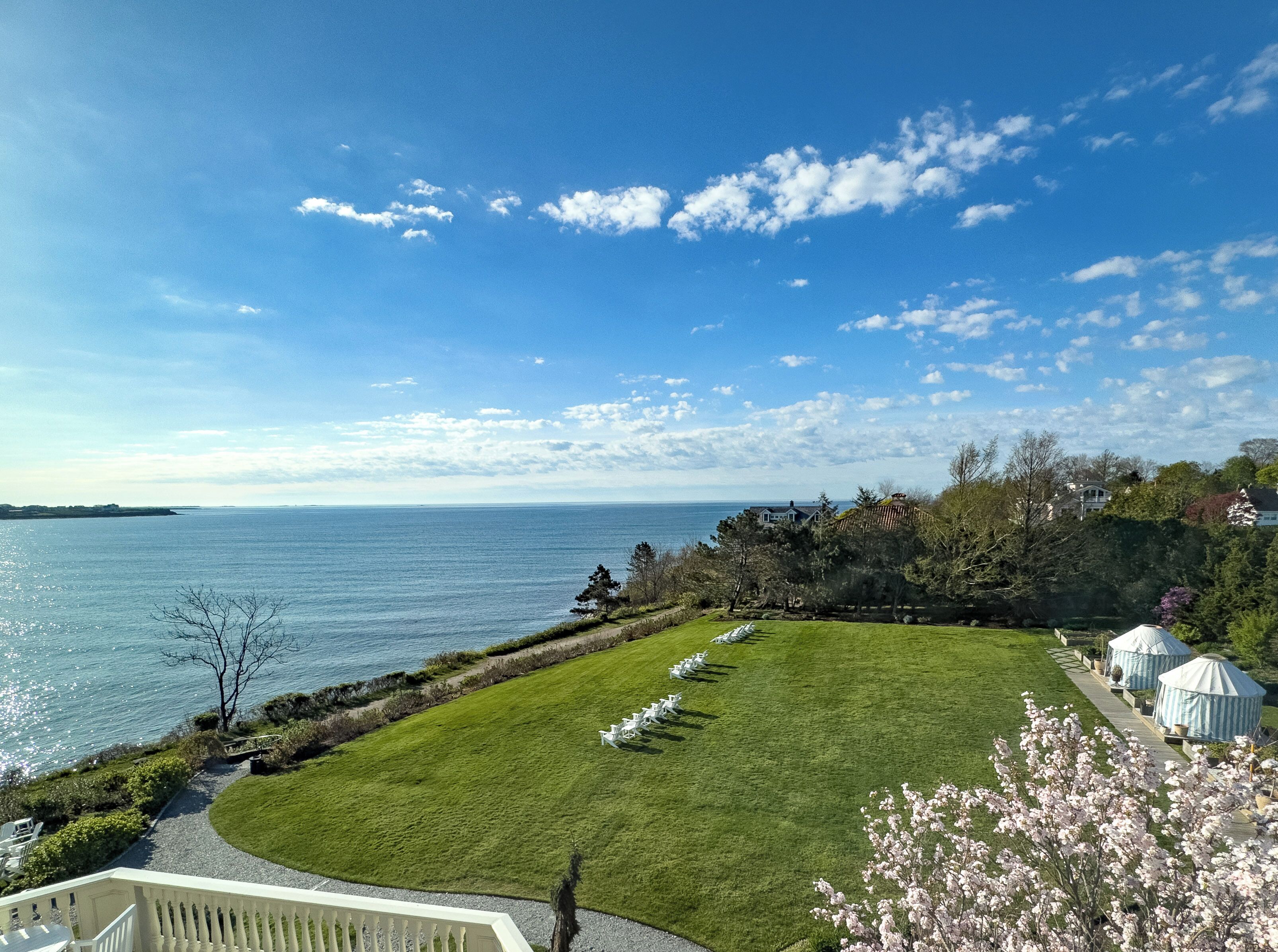 A coastal view with a grassy lawn, white chairs, and yurts by the water.